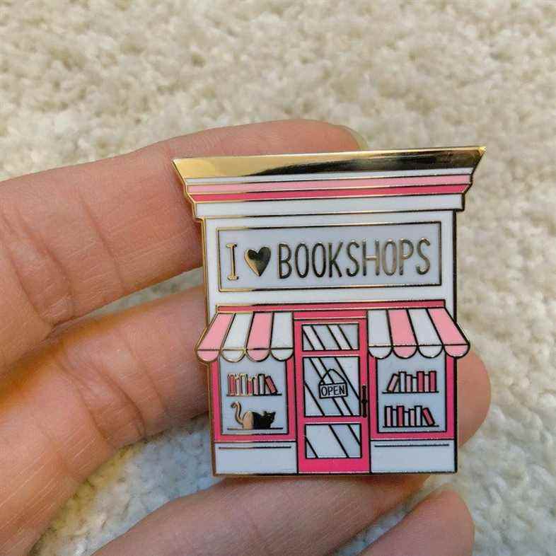 Image d'une épingle en émail.  C'est une devanture de librairie rose, avec les mots "I <3 librairies" en or. 