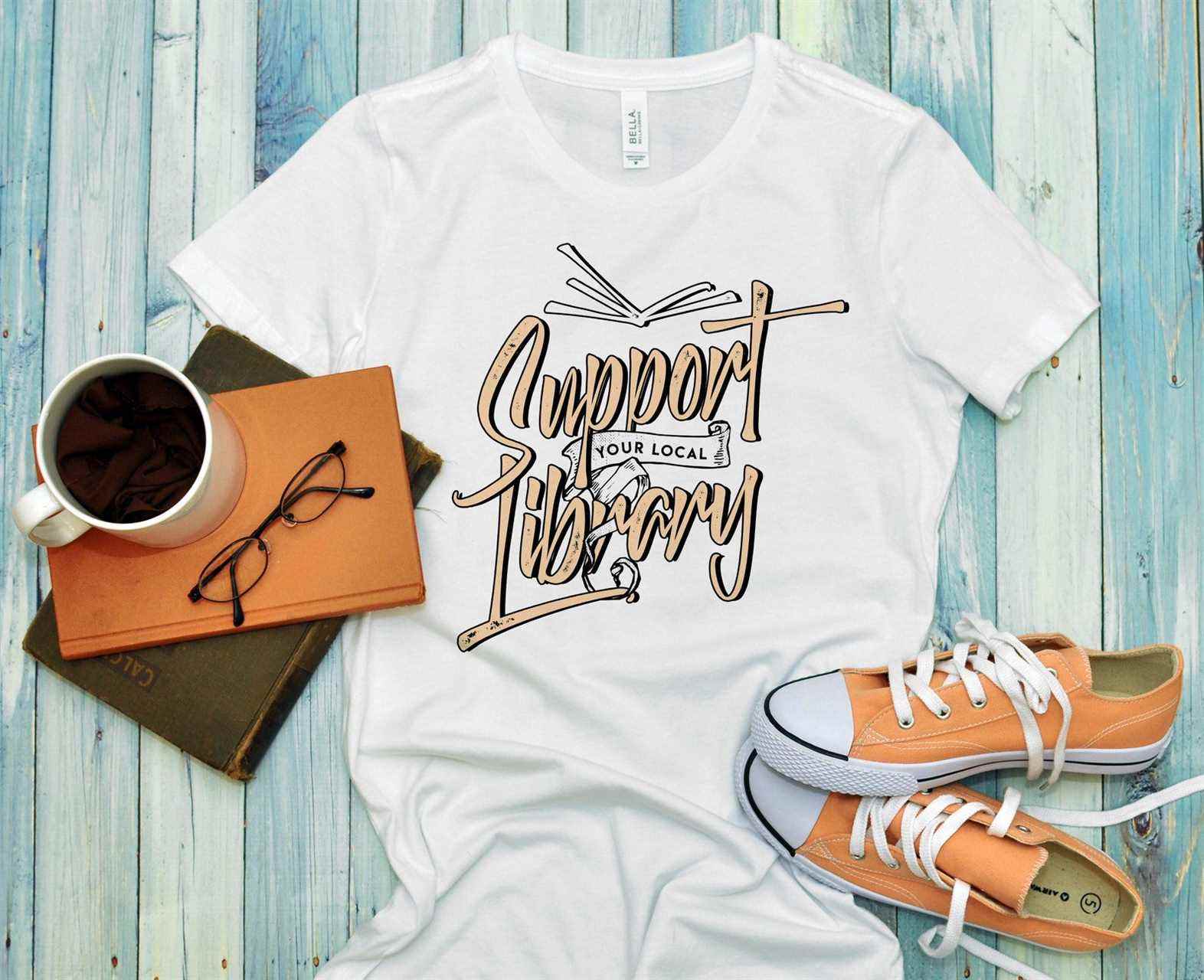 Image d'un t-shirt blanc qui indique "soutenez votre bibliothèque locale." C'est à côté d'une pile de livres et de chaussures orange. 