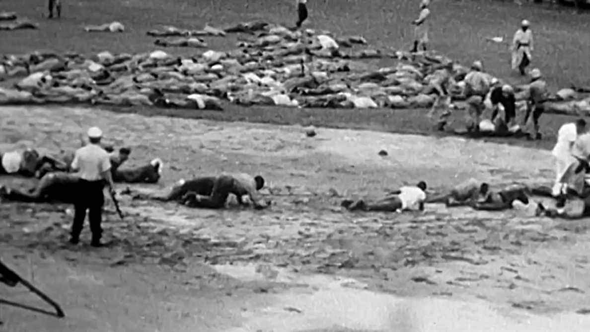 Une image vidéo en noir et blanc et granuleuse de dizaines d'hommes allongés sur le sol avec un garde armé se tenant au-dessus d'eux dans le documentaire Attica