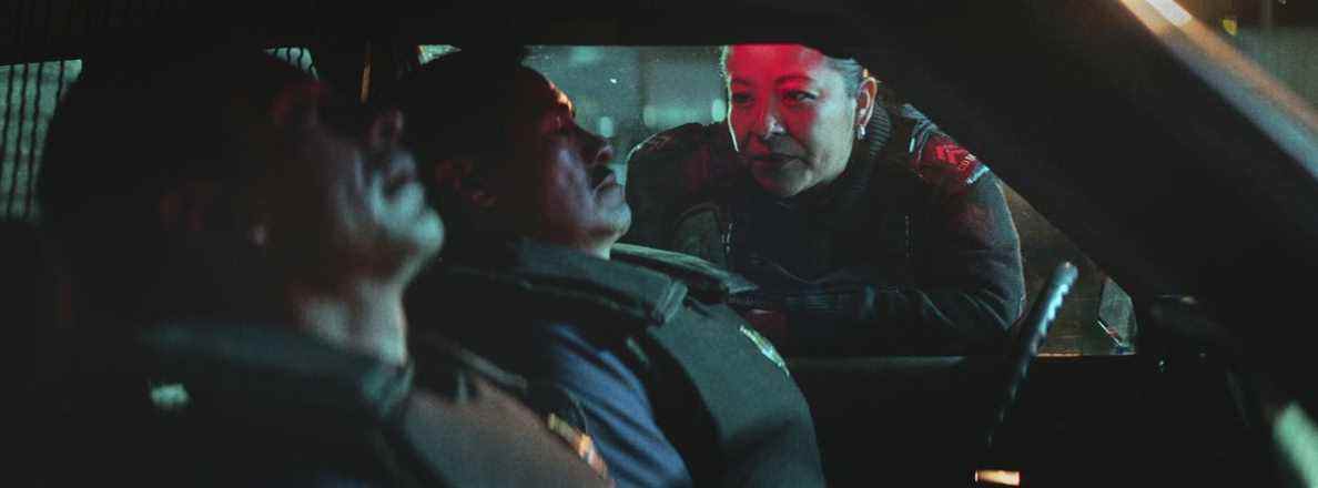 Une policière, le visage éclairé en rouge, se penche dans une voiture de flic aux teintes bleues dans A Cop Movie