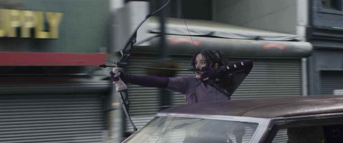 Kate Bishop se suspend à la fenêtre d'une voiture pour tirer une flèche sur un poursuivant lors d'une poursuite en voiture à Hawkeye