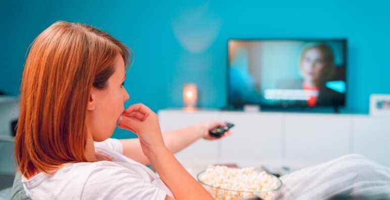 Young woman sitting on the bed sofa at home, lying with a popcorn bowl watching TV covered with blanket at her apartment alone enjoying movies or series holding remote control pointed