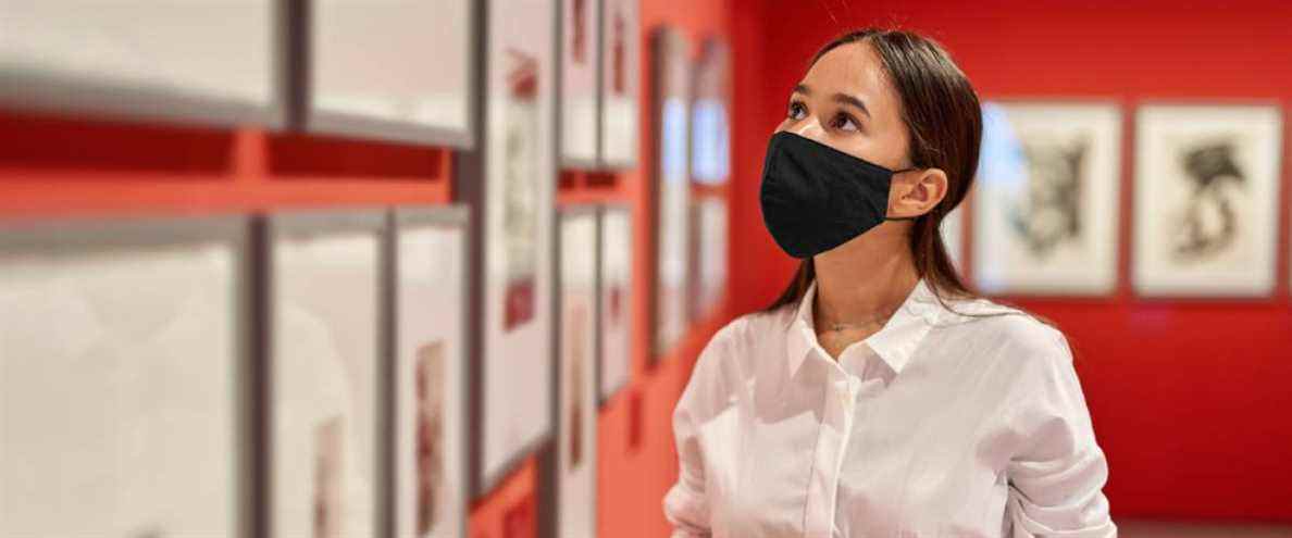 Une jeune femme portant un masque et tenant des lunettes de soleil vérifie des photos encadrées dans une galerie d'art