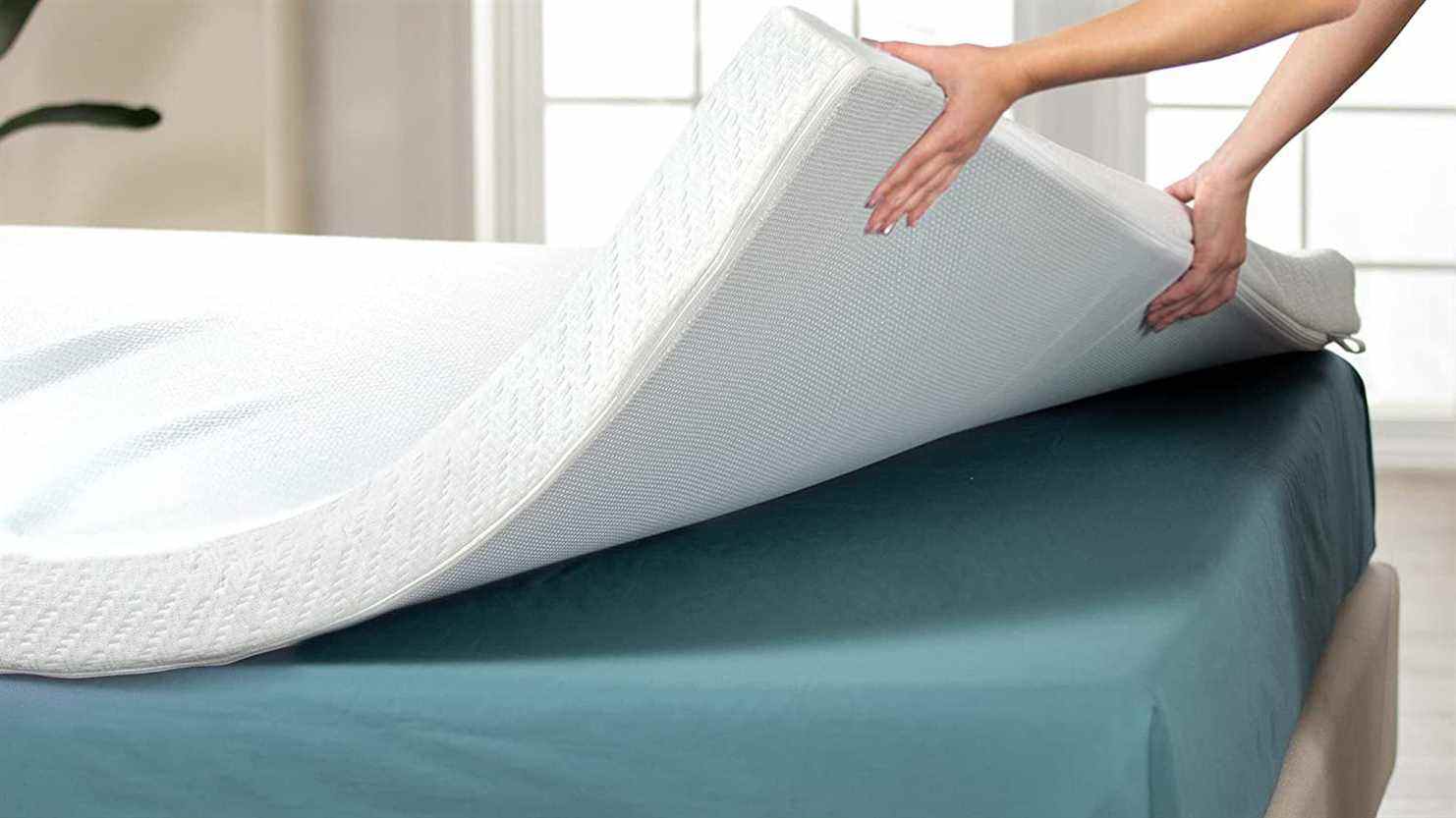 Une personne place un surmatelas blanc épais sur un matelas pour le rendre plus confortable pour dormir