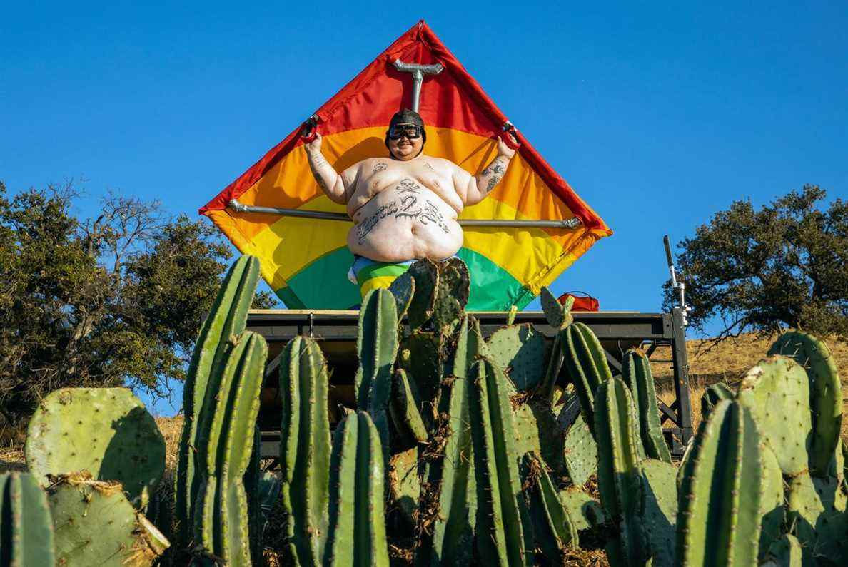 Zach Holmes, un homme tatoué extrêmement grand et torse nu, tient un deltaplane et sourit alors qu'il se tient au-dessus d'un champ de cactus
