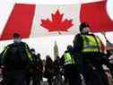 Des policiers passent devant un drapeau canadien géant devant le Parlement alors que les manifestants continuent de protester contre les mandats de vaccination vendredi à Ottawa.  L'Ontario a déclaré l'état d'urgence aujourd'hui à la suite des manifestations menées par des camionneurs qui paralysent la capitale et bloquent le commerce avec les États-Unis.