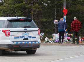 Des personnes en deuil rendent hommage devant le mémorial de fortune, construit à la mémoire des victimes de la fusillade de dimanche alors qu'un véhicule de la GRC passe à proximité à Portapique, en Nouvelle-Écosse, le 23 avril 2020.