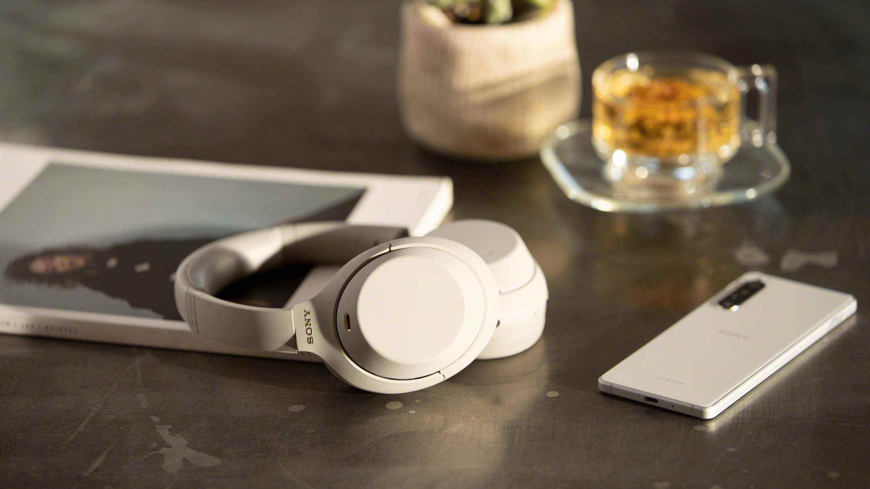Une paire d'écouteurs sony wh-1000xm4 en blanc sur une table sombre à côté d'un magazine et d'un smartphone argenté