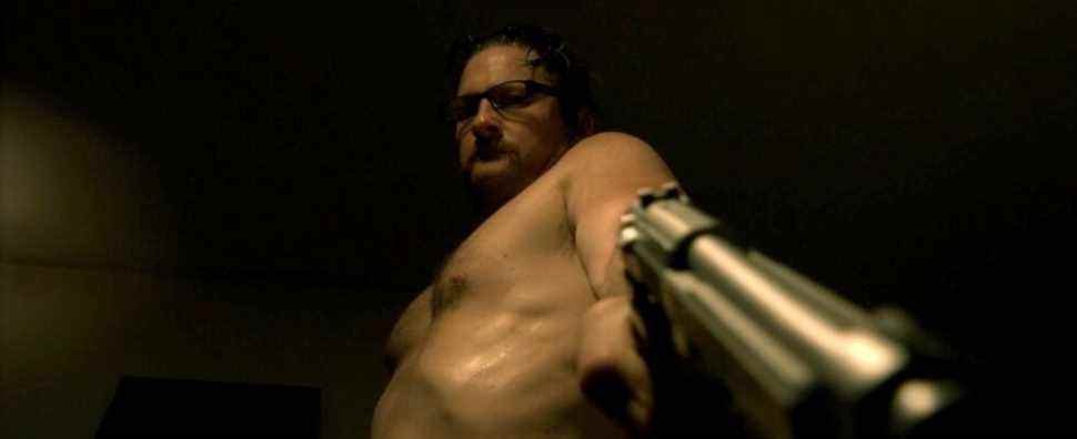A shirtless Ray Belfi points a gun downward