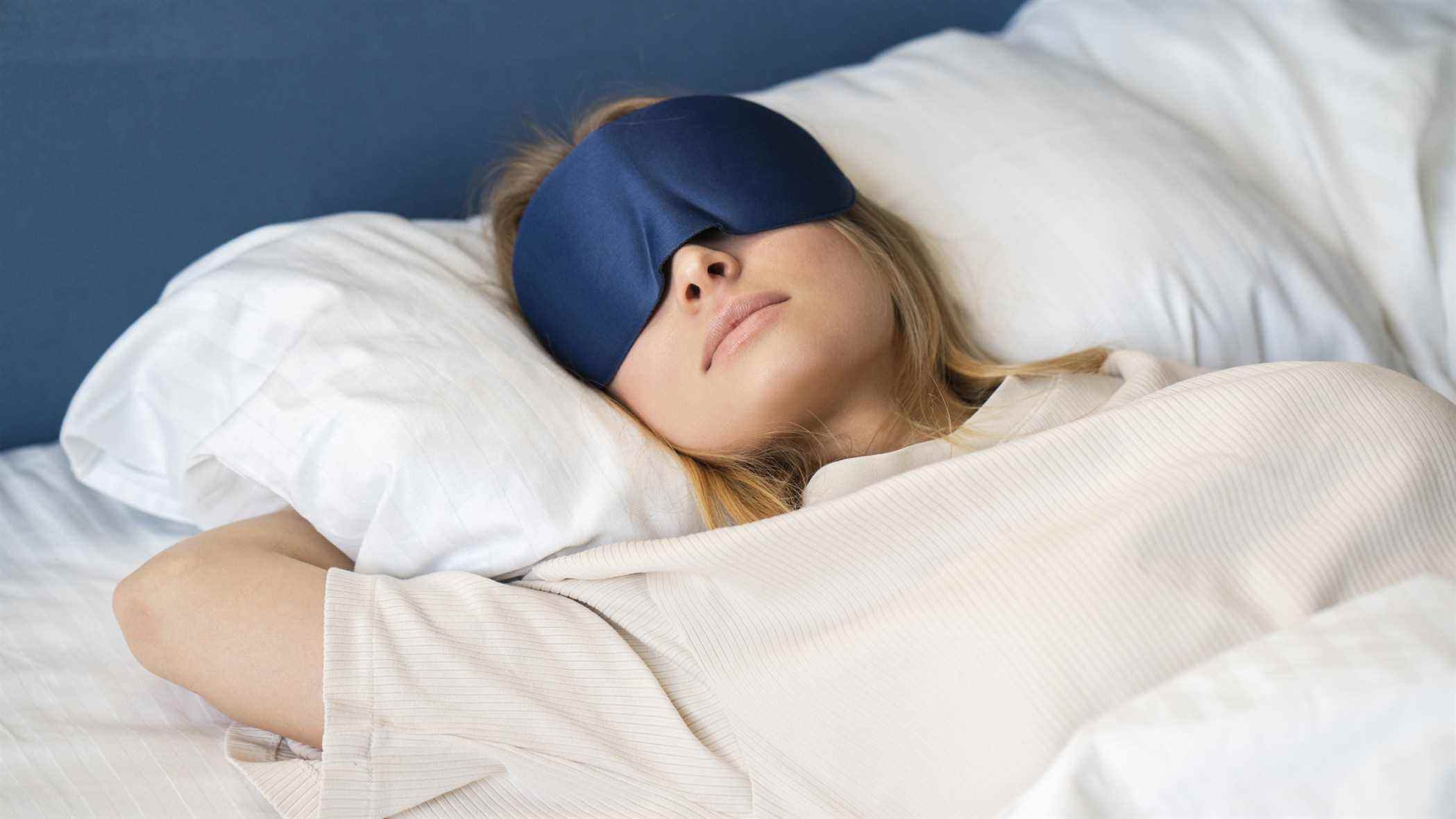 Une femme aux cheveux blonds portant un masque bleu pour les yeux dort sur un canapé-lit