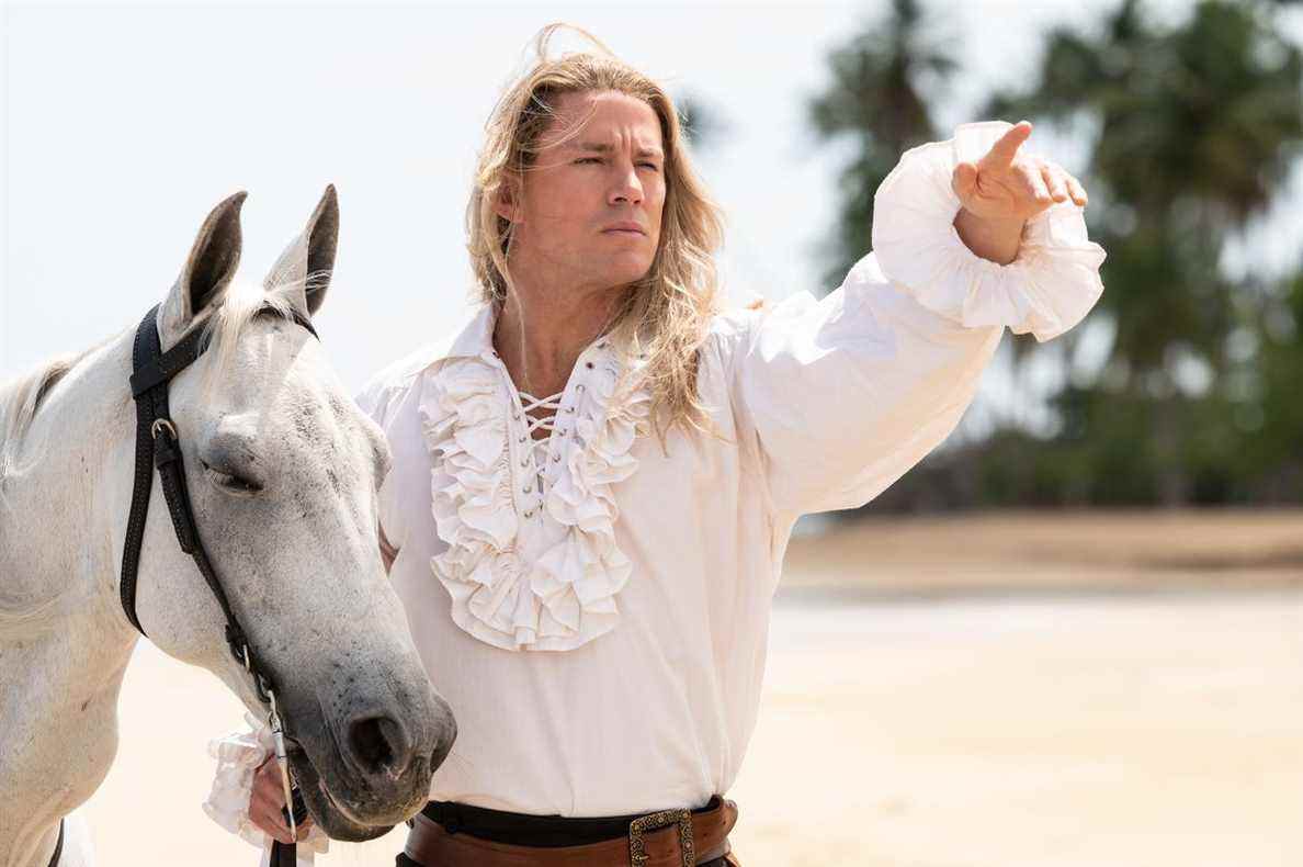 Channing Tatum, dans une chemise blanche à froufrous, pointe au loin tout en se tenant debout avec un cheval blanc sur une plage de The Lost City