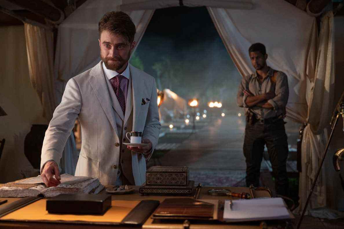 Daniel Radcliffe en costume blanc tient une tasse de thé et se tient au-dessus d'un bureau dans une tente dans The Lost City