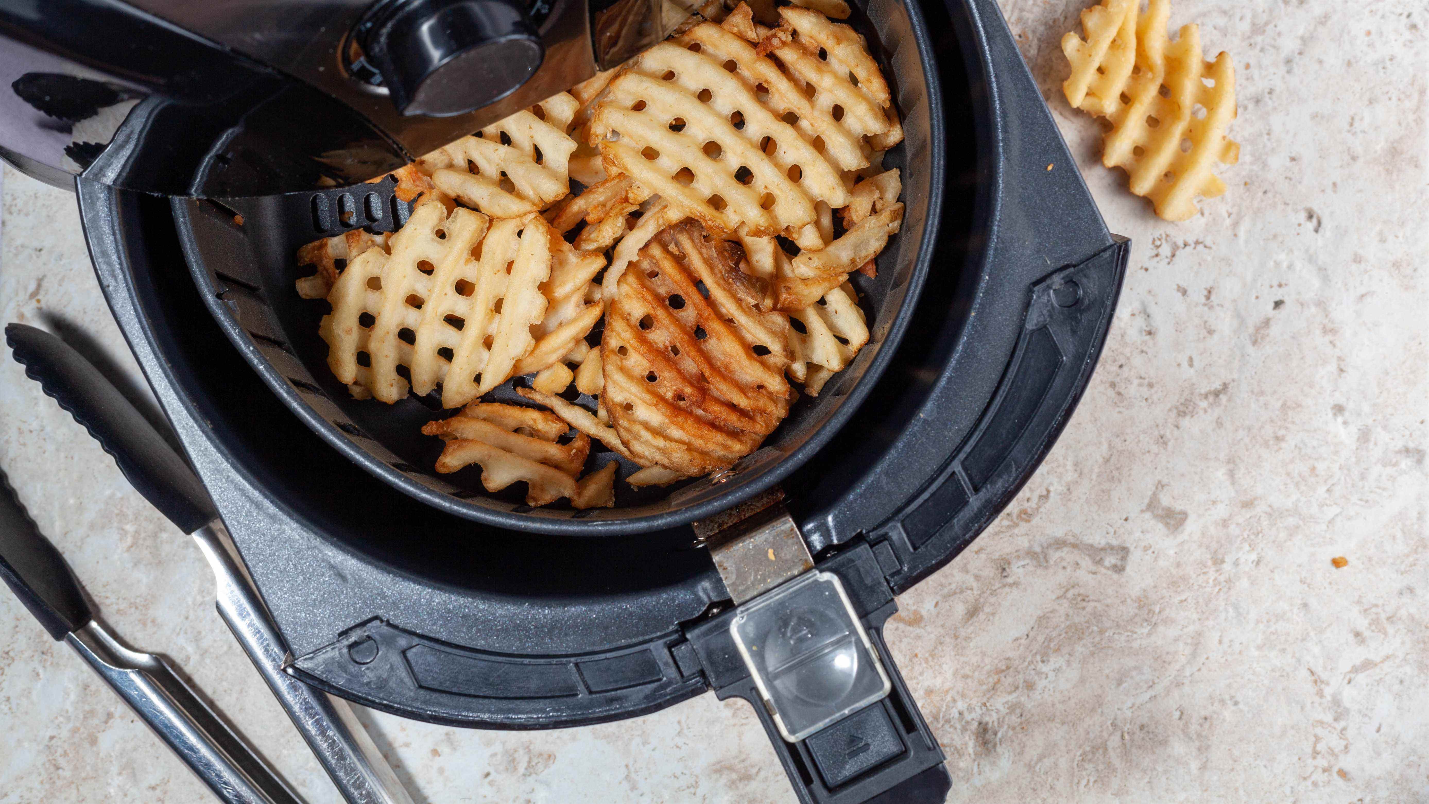 Un panier de friteuse à air avec des gaufres de pommes de terre à côté de pinces en silicone