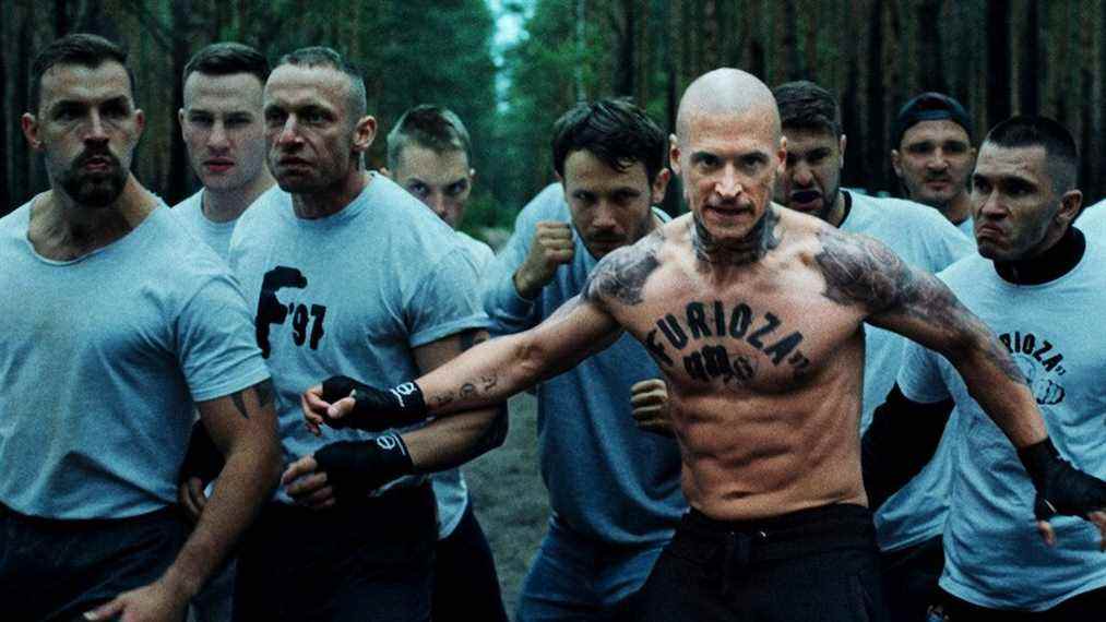 Un homme avec le mot 'Furioza' tatoué sur sa poitrine se tient devant un groupe d'hommes portant des chemises grises identiques à Furioza.