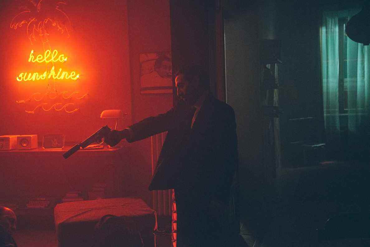 Marcello Fonte pointant un pistolet dans une pièce sombre éclairée par une enseigne au néon dans The Turning Point (2021).