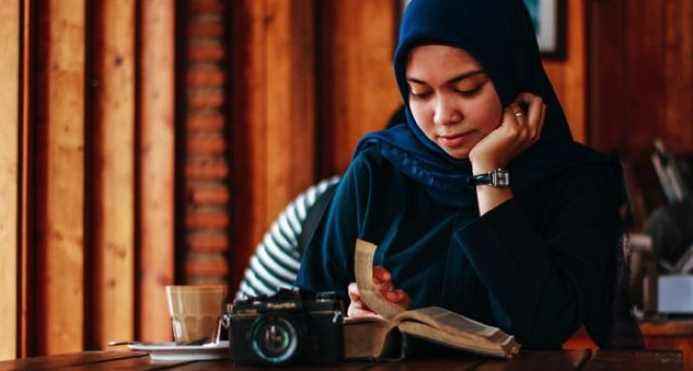 femme à la peau bronzée claire lisant en hijab, appareil photo et livre devant elle sur une table