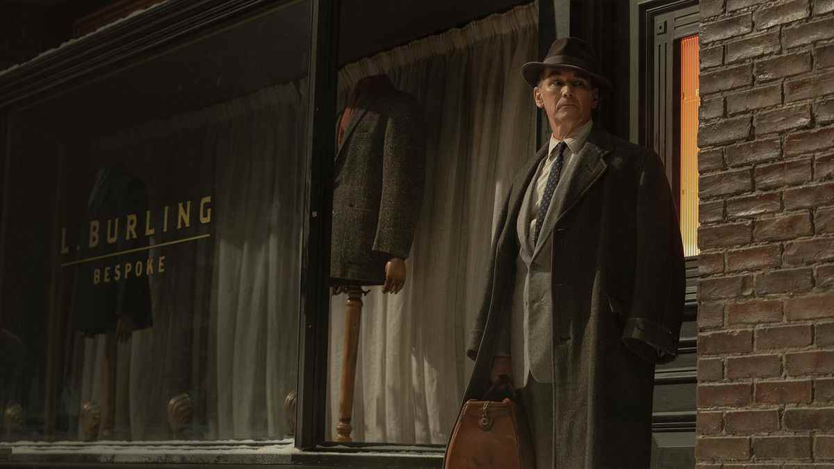Mark Rylance dans le rôle de Leonard en costume et manteau devant sa boutique de tailleur du coin dans The Outfit (2022).