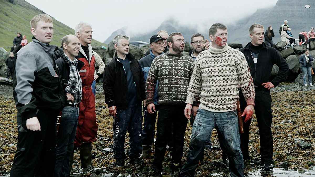 Un groupe d'hommes en sweat-shirts, dont l'un est couvert de sang, se tient sur le rivage d'une plage dans A Taste of Whale.
