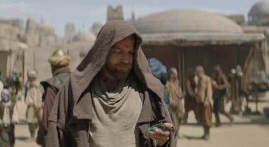 A middle-aged man with sandy hair and beard, wearing a brown hooded cloak and looking down at something in his hand as he stands in a desert city; still from "Obi-Wan Kenobi."