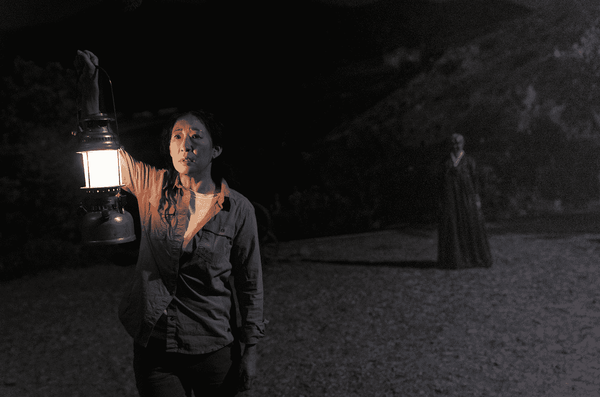 Sandra Oh dans le rôle de Beth tenant une lanterne à l'extérieur alors qu'une silhouette inquiétante se tient derrière eux à Umma.