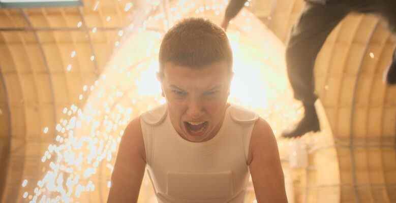 Medium-close shot of a child wearing a white vest and screaming while sparks fly and a figure is thrown backward behind her; still of Millie Bobby Brown in "Stranger Things 4."