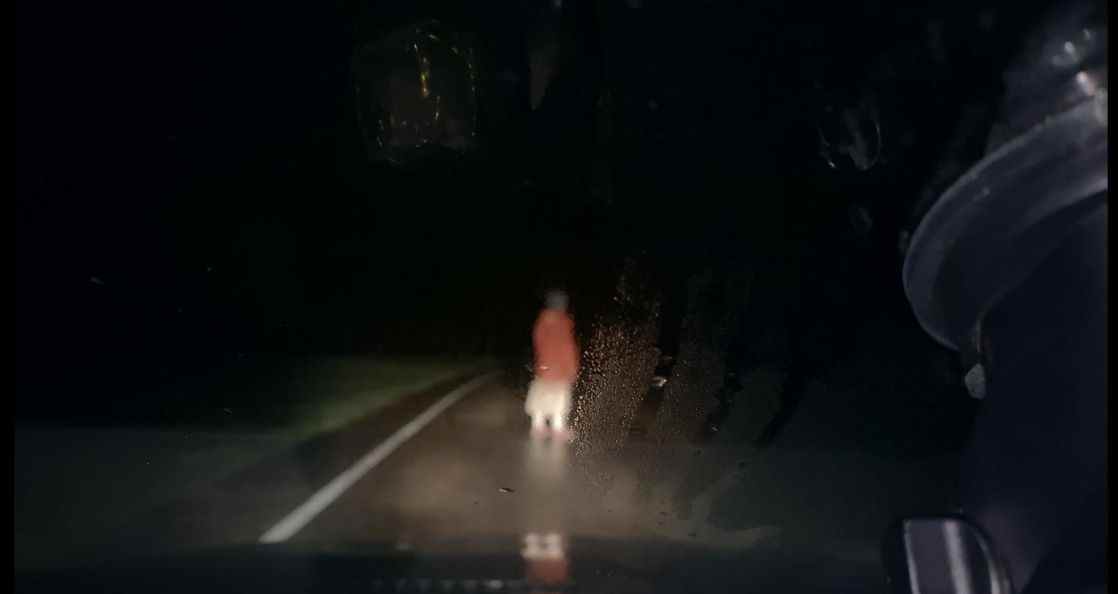 Une femme se tient au milieu d'une route la nuit éclairée par les phares d'une voiture en Dashcam.