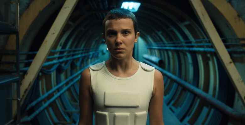 A teen girl with short buzzed hair wearing a white protective vest enters a mechanical hallway; still from "Stranger Things 4."