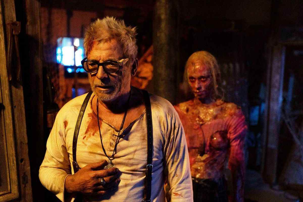Un homme échevelé portant des lunettes avec du sang sur sa chemise serre un pendentif autour de son cou avec une silhouette macabre ressemblant à un zombie en arrière-plan.