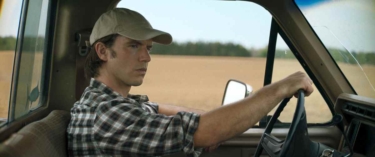 Un jeune homme vêtu d'une chemise en flanelle à carreaux et d'une casquette de baseball est assis au volant d'un camion à plateau tout en passant devant une plaine de terre.
