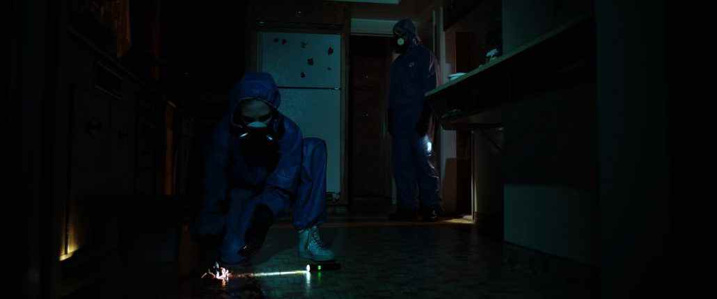 Deux personnages en tenue de protection contre les matières dangereuses inspectent le sol d'une cuisine sombre dans 11th Hour Cleaning.