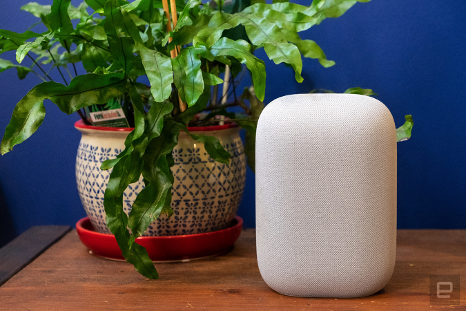 Haut-parleur intelligent Google Nest Audio posé sur une tablette en bois à côté d'une plante verte, contre un mur bleu.