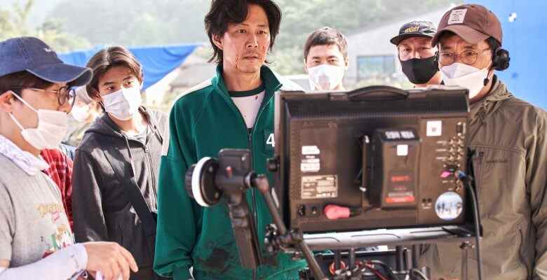 A man in a dark green tracksuit examines a television monitor, surrounded by others in masks; actor Lee Jung-jae behind the scenes of "Squid Game."