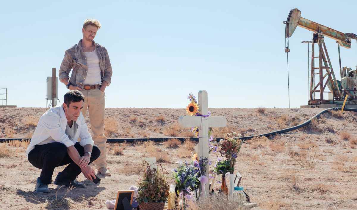 BJ Novak et Boyd Holbrook se tiennent près d'une croix commémorative parsemée de bouquets dans un champ jaune aride près d'un derrick de pétrole à Vengeance