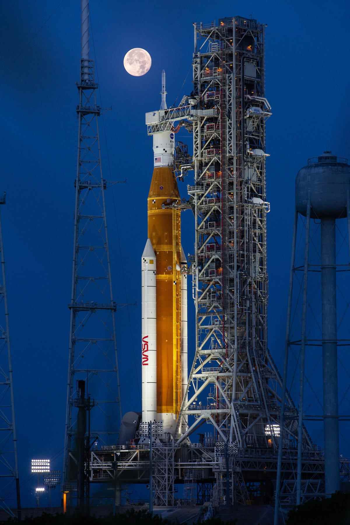 Sur un ciel bleu nuit, une pleine lune est visible vers le haut à gauche de l'image et la fusée orange Artemis I de la NASA et le vaisseau spatial Orion sont installés au premier plan.