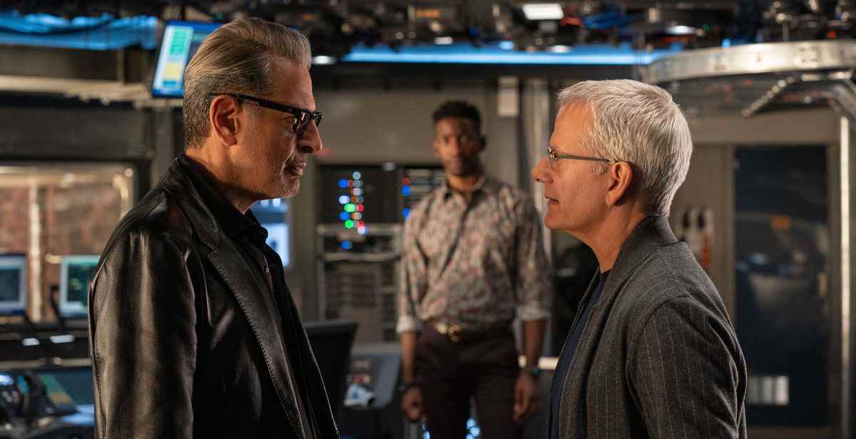 Le Dr Ian Malcolm (Jeff Goldblum) et Lewis Dodgson (Campbell Scott) s'affrontent au premier plan d'une salle à haute technologie, de style flingueur, tandis que Ramsay Cole (Mamoudou Athie) regarde en arrière-plan dans Jurassic World Dominion.