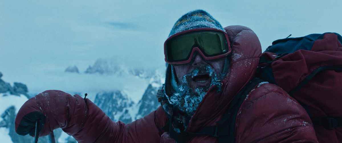 Un homme en équipement d'alpinisme rouge est couvert de neige, en particulier sa barbe.  Il porte un sac à dos rouge et regarde au-dessus de la caméra, avec des montagnes enneigées derrière lui.