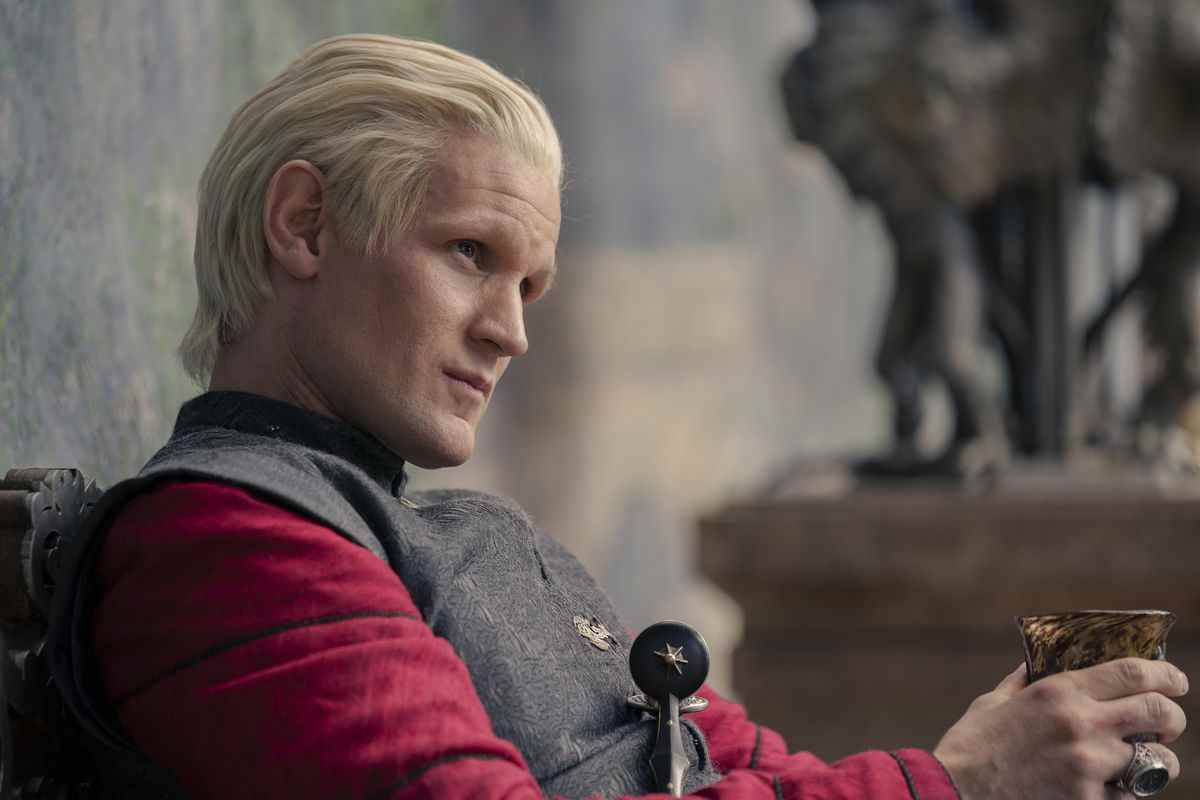 Daemon Targaryen, graissé, vêtu d'une chemise rouge et d'une robe grise, fixant quelqu'un alors qu'il serre son vin dans House of the Dragon