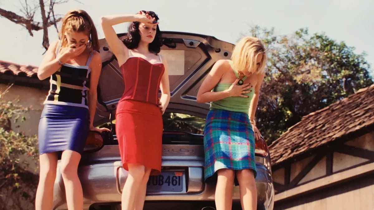 trois adolescentes aux couleurs vives se tiennent à côté d'un coffre de voiture ouvert ;  deux d'entre eux bâillonnent;  celui du centre, qui porte du rouge vif, a l'air calme et presque ennuyé