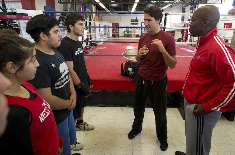 Un homme en T-shirt bordeaux discute avec trois jeunes, un ring de boxe derrière lui et un entraîneur à côté de lui.