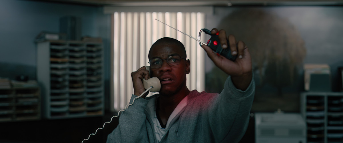 John Boyega porte un sweat à capuche gris et tient un détonateur dans une main pendant qu'il est au téléphone dans l'autre dans Breaking.