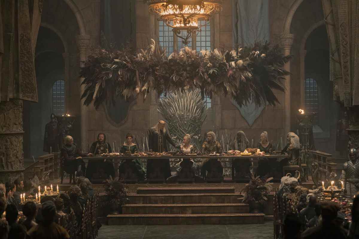 Une photo de la table royale dans la grande salle de King's Landing, avec (de gauche à droite) : Daemon, Ser Strong, Alicent, Viserys, Rhaenyra, Laenor, Corlys, Rhaenys et Laena autour d'elle face à d'autres tables en dessous d'eux.  Viserys est debout et regarde Rhaenyra avec son bras sur son épaule.