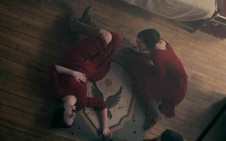 Deux femmes sur le sol d'une pièce, portant des tuniques rouges et des bonnets noirs.  Les deux saignent de la bouche et l'un d'eux est allongé inconscient sur le sol;  encore de 'The Handmaid's Tale."