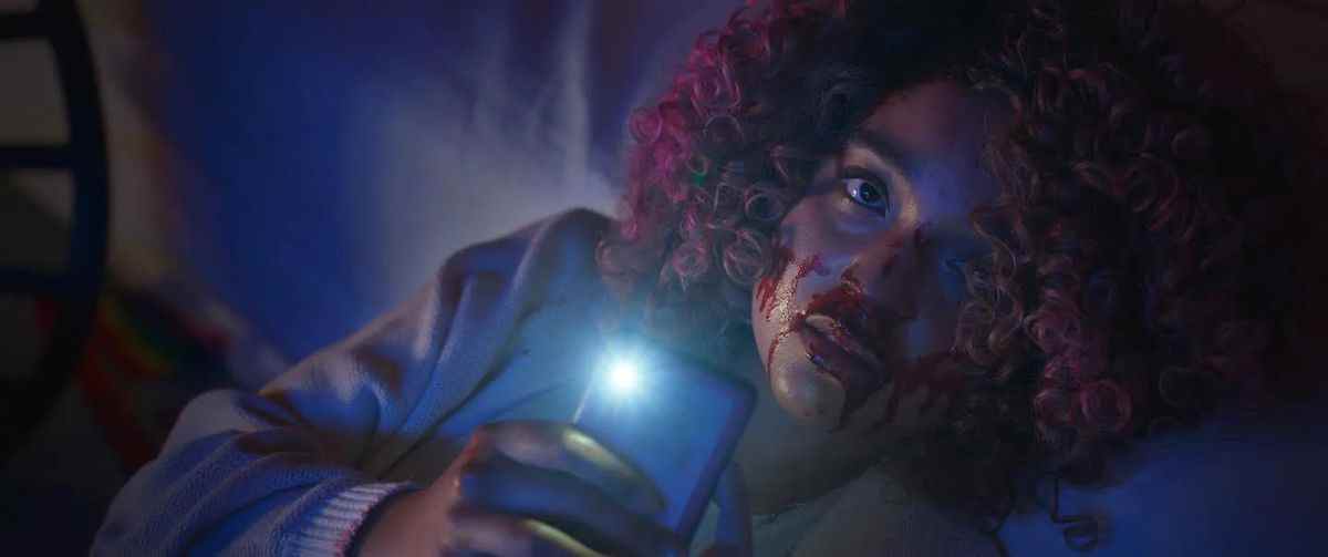 Une femme avec du sang coulant sur son nez et son visage avec des boucles afro roses tient un téléphone portable dans sa main avec la lumière de l'appareil photo allumée.