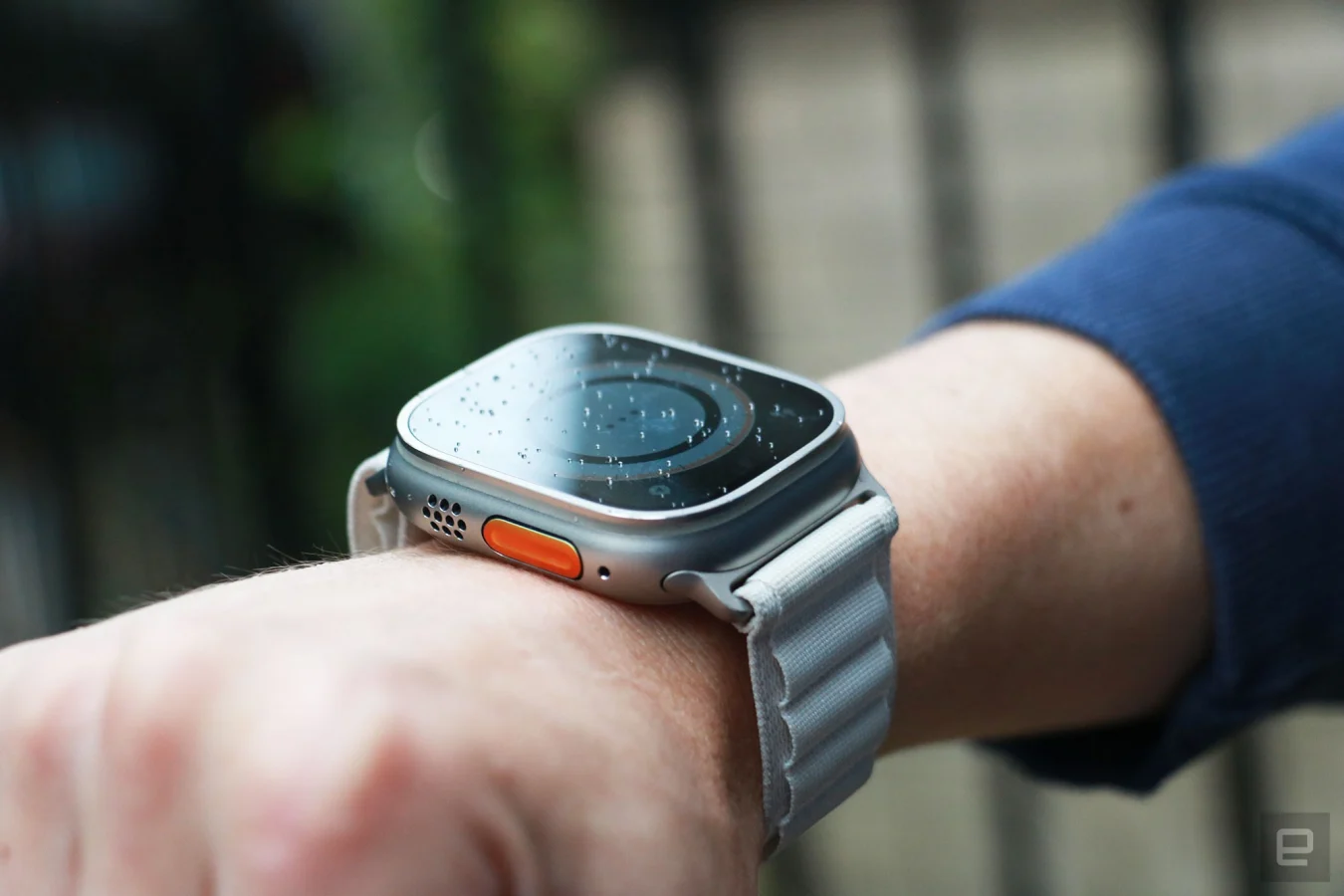 Vue hors angle de l'Apple Watch Ultra avec boucle alpine sur le poignet d'une personne.  De ce point de vue, le bouton Action orange et la grille du haut-parleur sur le bord gauche sont visibles, tandis que le contenu de l'écran est sombre.