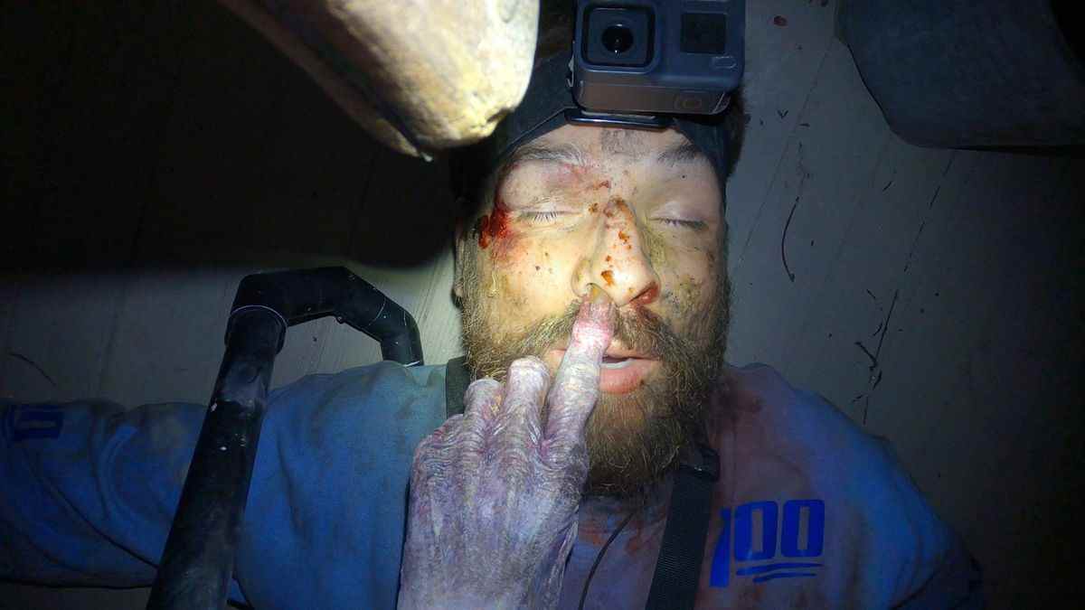 Un homme portant une caméra frontale est allongé inconscient sur un plancher en bois alors qu'une main zombifiée enfonce son doigt dans l'une de ses narines.