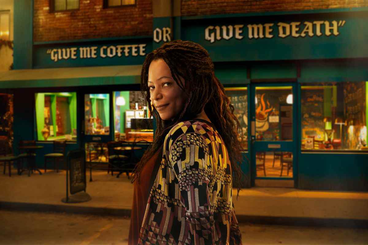 une femme avec des dreadlocks sombres sourit devant un magasin qui dit DONNEZ-MOI DU CAFÉ OU DONNEZ-MOI LA MORT
