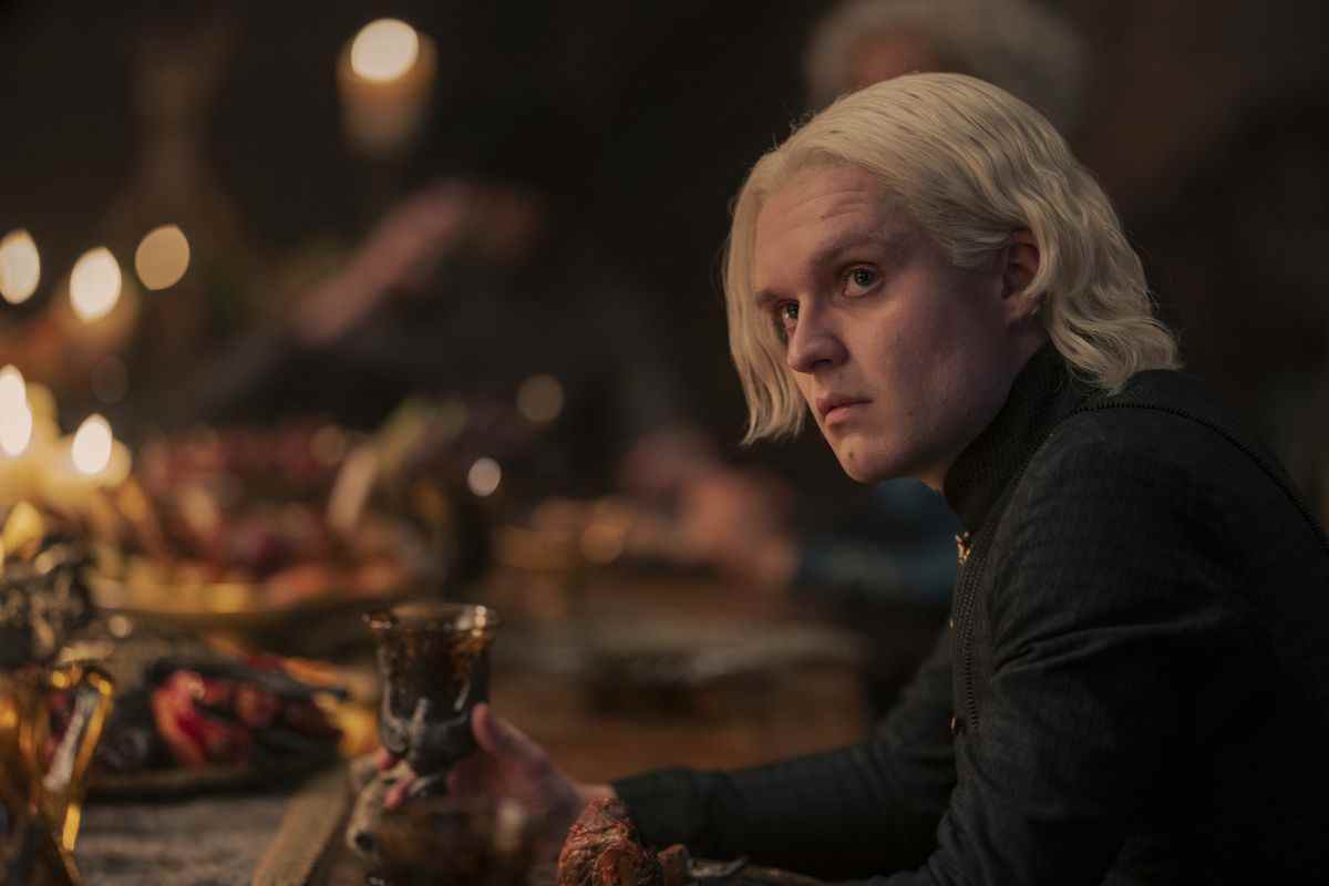 Tom Glynn-Carry dans le rôle d'Aegon II Targaryen assis à une table avec un festin tenant une coupe gagnante dans House of the Dragon