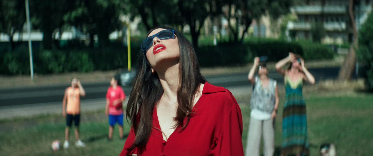 Une femme en rouge porte des lunettes noires et regarde le soleil, alors que les gens derrière elle dans un parc lèvent également les yeux, dans Dark Glasses.