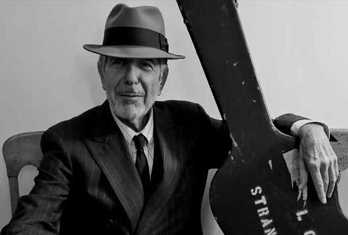 Leonard Cohen, vêtu d'un costume-cravate et d'un joli chapeau, tient un étui à guitare.