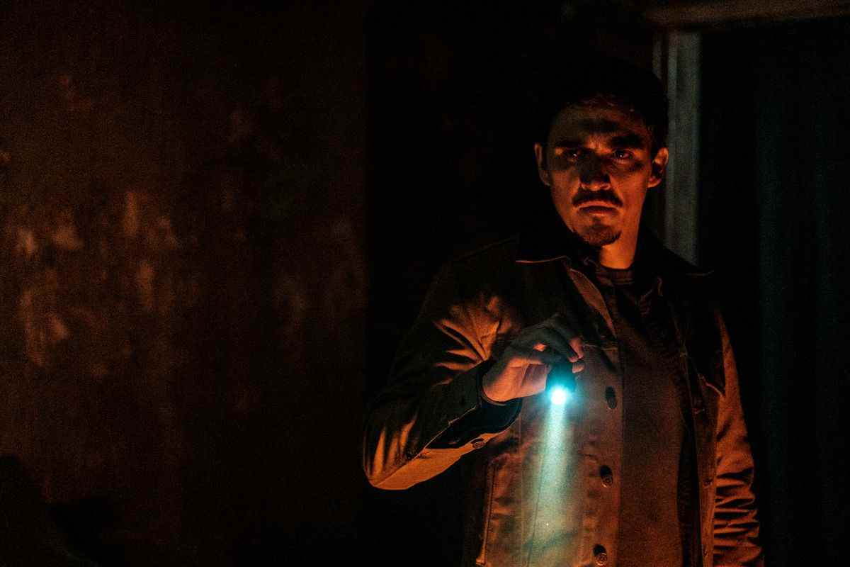 Joel (Kyle Gallner) se tient dans une pièce sombre et éclairée en rouge tenant une lampe de poche bleu vif dans Smile